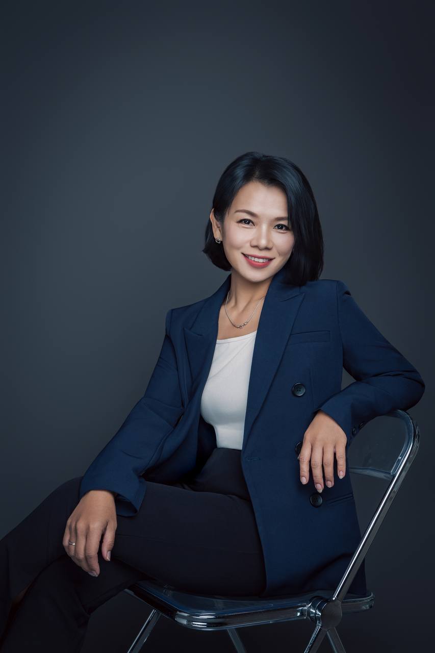 宇宙媽媽賞身穿深藍色西裝外套，側坐在透明椅上，手靠椅背微笑
